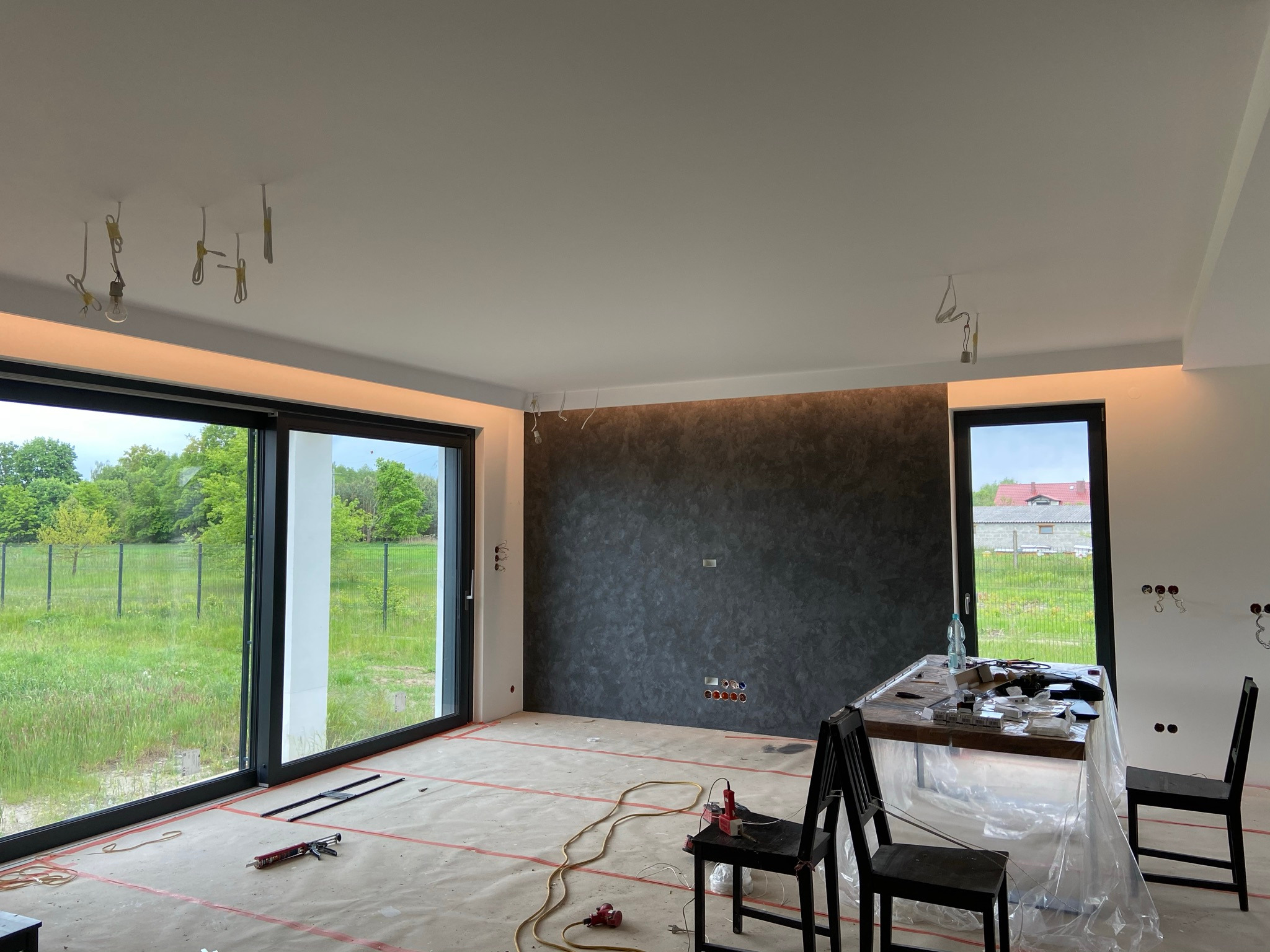 Wnętrze w trakcie wykańczania: widoczne wyprowadzenia elektryczne na suficie, narzędzia na podłodze zabezpieczonej folią, oraz stół z akcesoriami elektrycznymi pod ścianą z dekoracyjnym tynkiem.