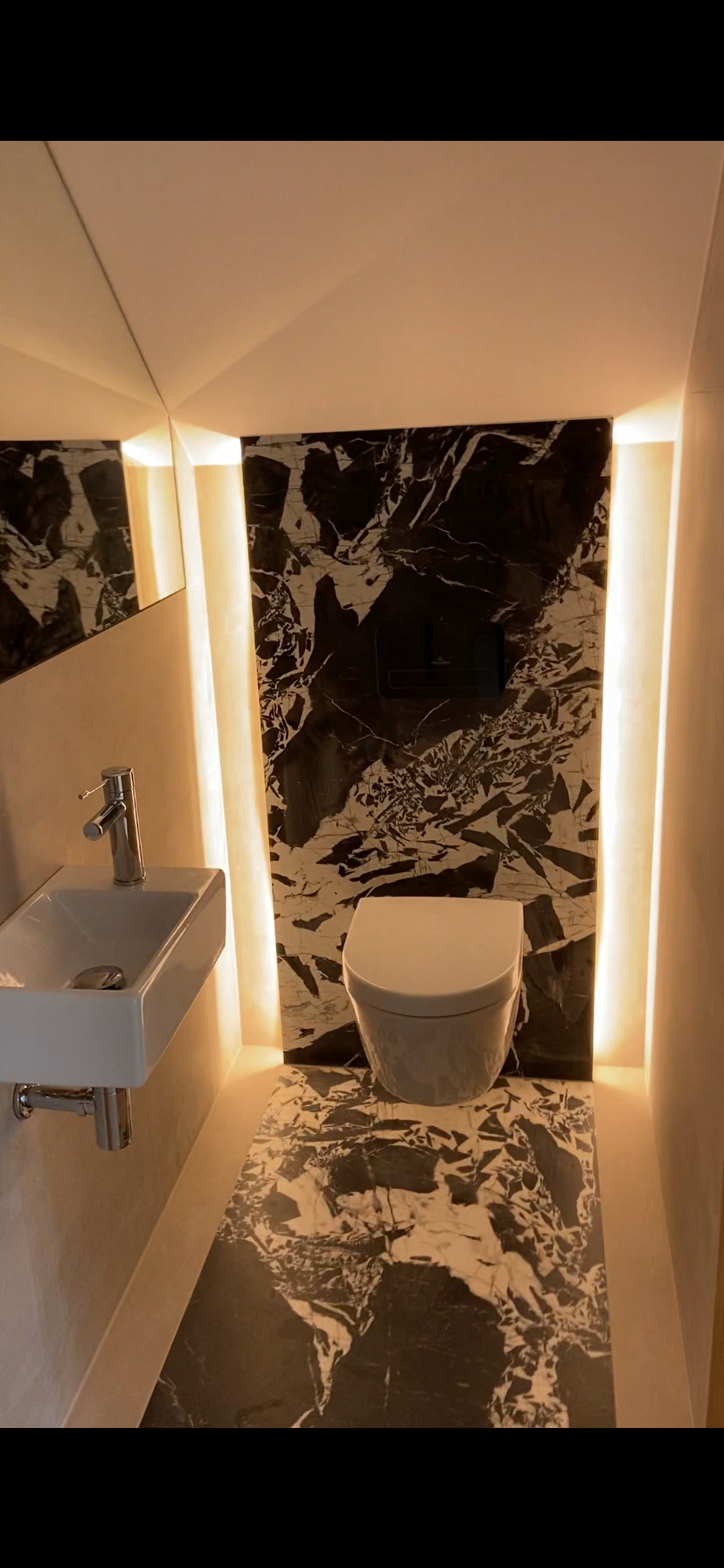 Nowoczesna, mała łazienka z czarno-białym marmurem na ścianie i podłodze, podświetlana ciepłym światłem, z wiszącą miską WC i umywalką.