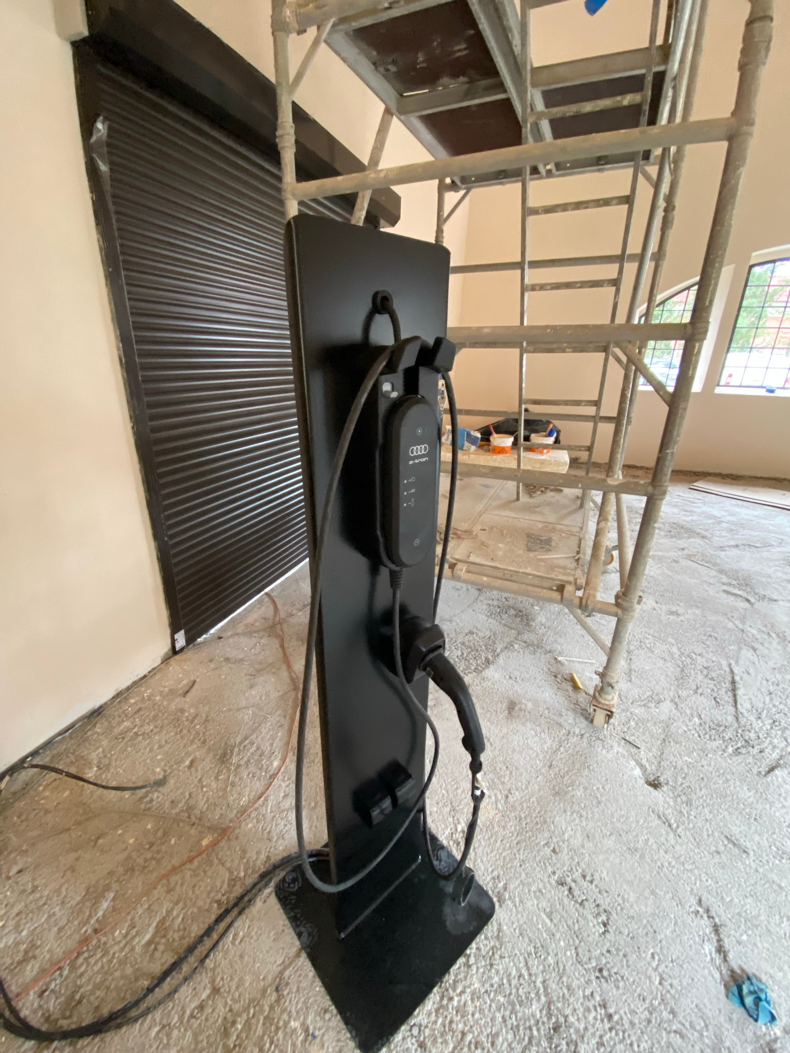 Czarna ładowarka do samochodu elektrycznego marki Audi e-tron, umieszczona na tle surowego wnętrza w trakcie remontu, widoczne rusztowanie i brama garażowa z roletą.