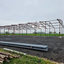 Szkielet konstrukcji stalowej hali w trakcie budowy, widoczne elementy konstrukcyjne i przygotowane materiały budowlane na placu budowy.