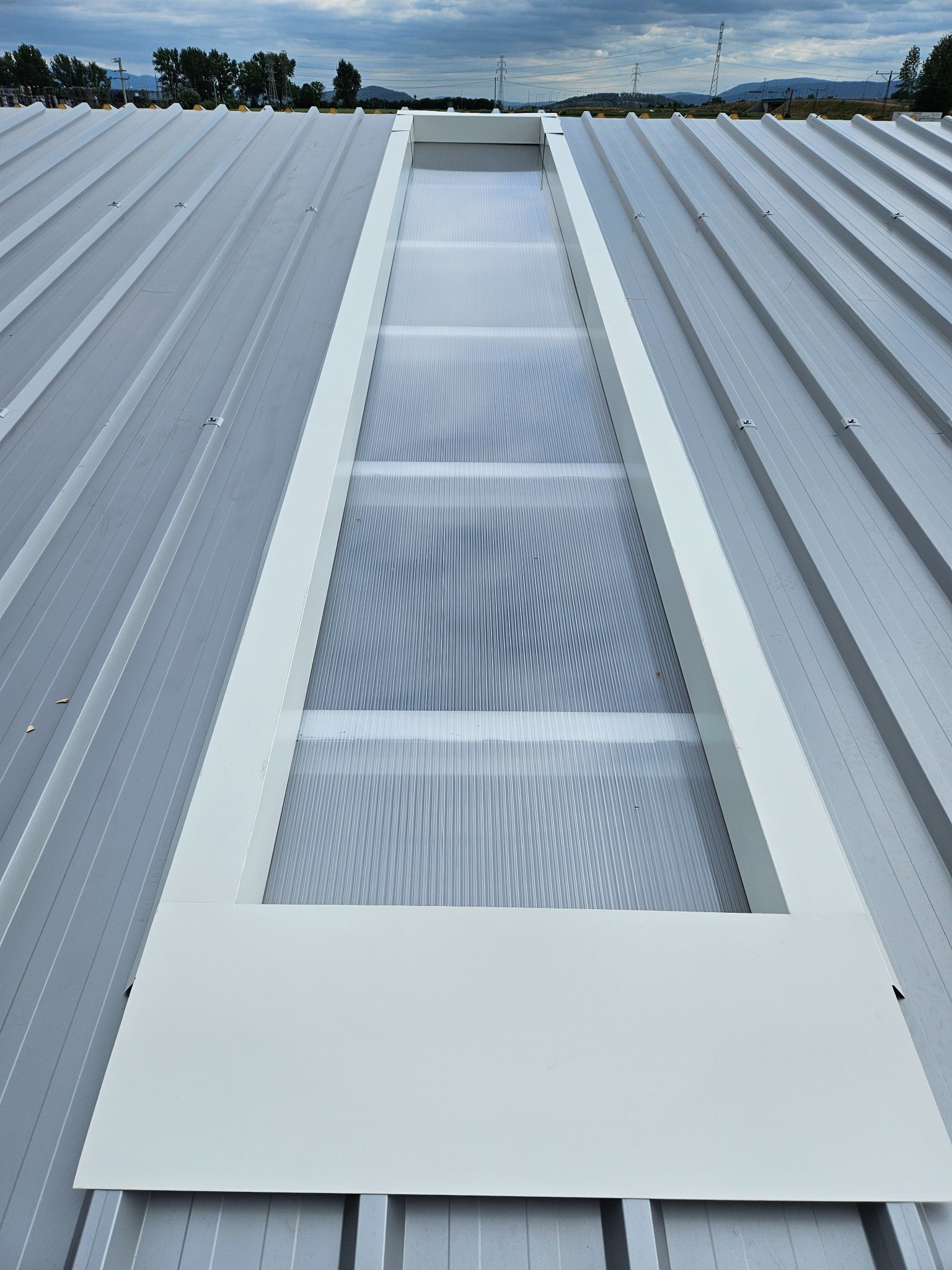 Widok z góry na metalowy dach z wbudowanym świetlikiem z poliwęglanu w białej ramie, widoczne mocowania blachy, w tle zieleń i góry pod pochmurnym niebem.