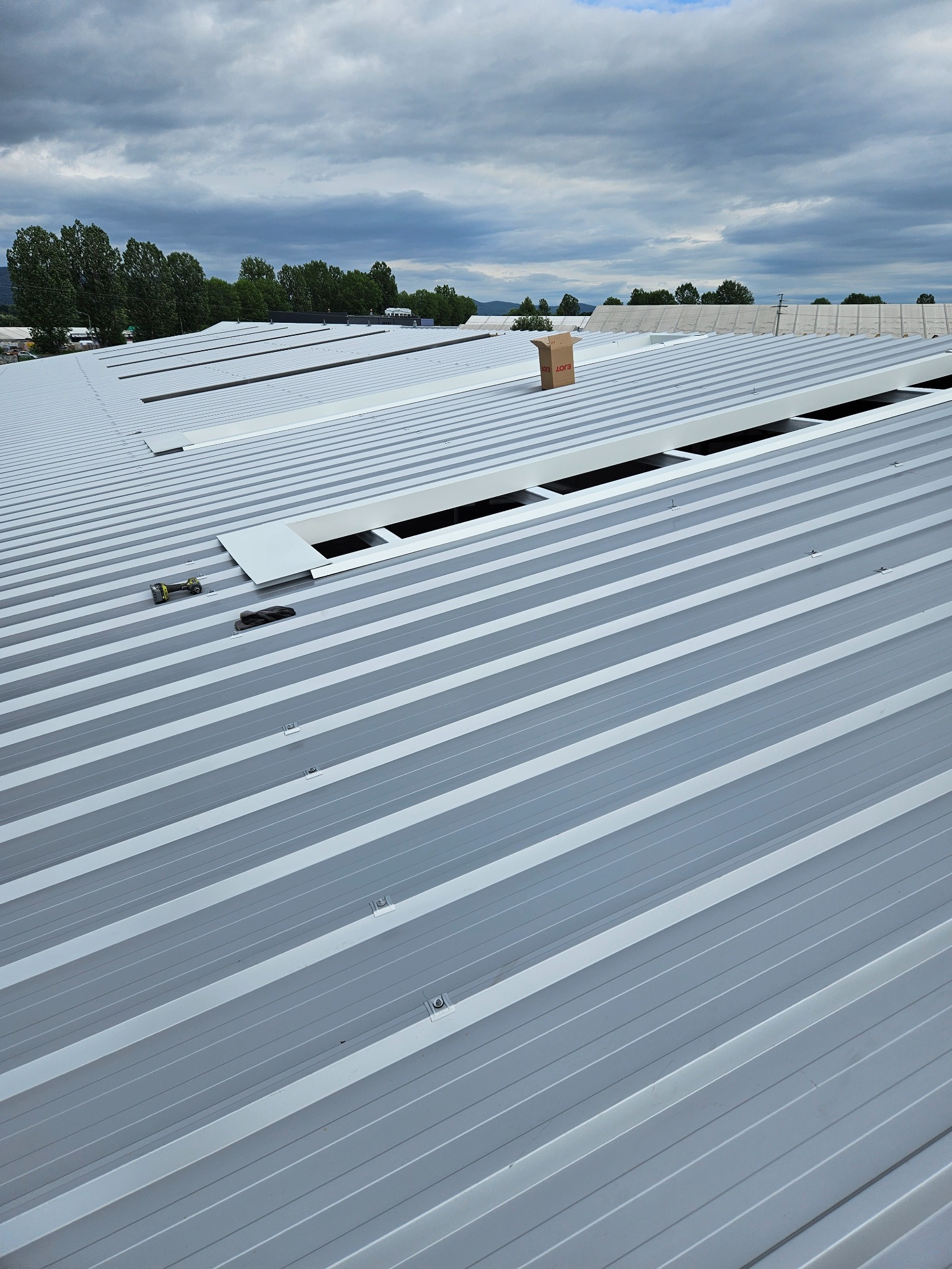 Widok z góry na połacie dachu hali przemysłowej z płyt warstwowych, z otworami okiennymi, narzędziami i kartonem na powierzchni, w tle drzewa i pochmurne niebo.