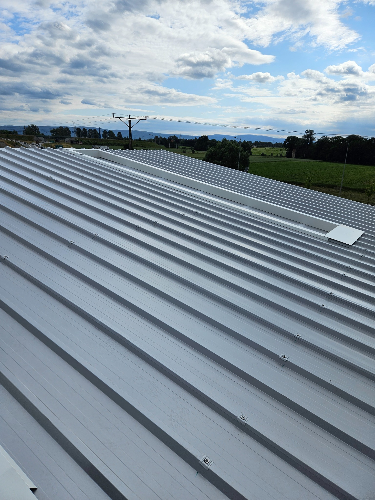 Szary dach z płyt warstwowych z widocznymi mocowaniami, na tle błękitne niebo z chmurami i odległy krajobraz.