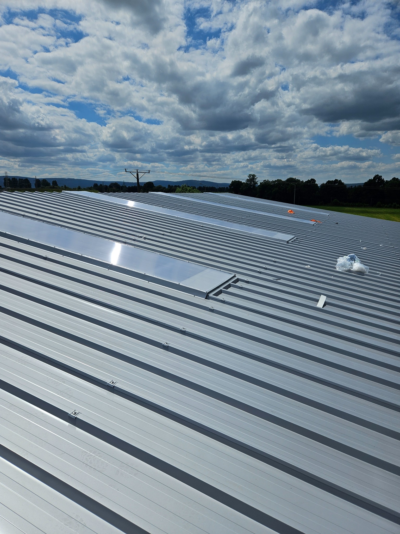 Nowo zamontowany, srebrny dach z blachy trapezowej z świetlikami, widok z góry na tle błękitnego nieba z chmurami i odległego krajobrazu.