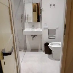 Widok na wyremontowaną łazienkę z białymi płytkami, umywalką, toaletą i uchwytami dla niepełnosprawnych, widzianą zza uchylonych drzwi z widoczną podłogą z desek przed wejściem.