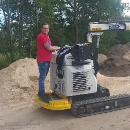 Operator w okularach stoi na platformie minikoparki gąsienicowej Palfinger TGT Robby 2000 na tle hałd piasku i drzew.