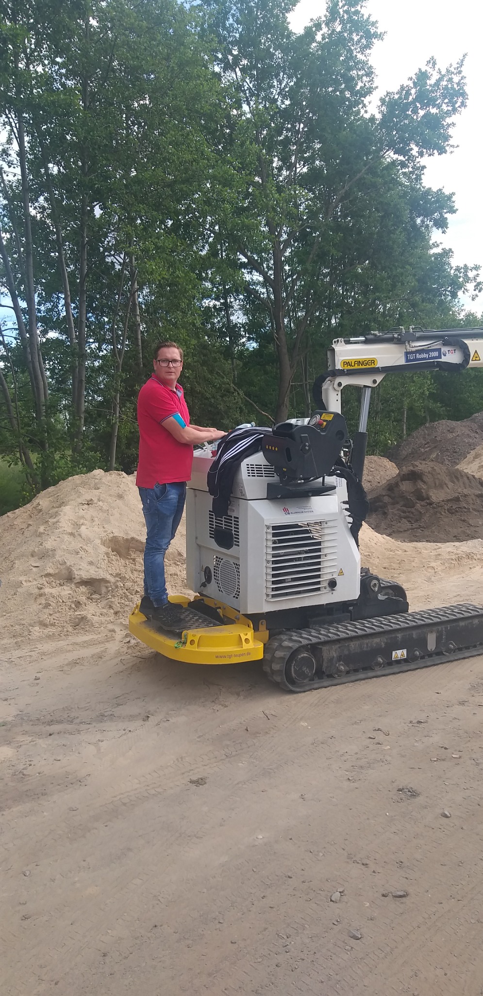 Operator w okularach stoi na platformie minikoparki gąsienicowej Palfinger TGT Robby 2000 na tle hałd piasku i drzew.