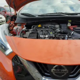 Otwarta maska pomarańczowego samochodu Nissan Micra, widoczny silnik, zbiornik płynu chłodniczego i akumulator. W tle zaparkowane inne pojazdy na żwirowym podłożu.