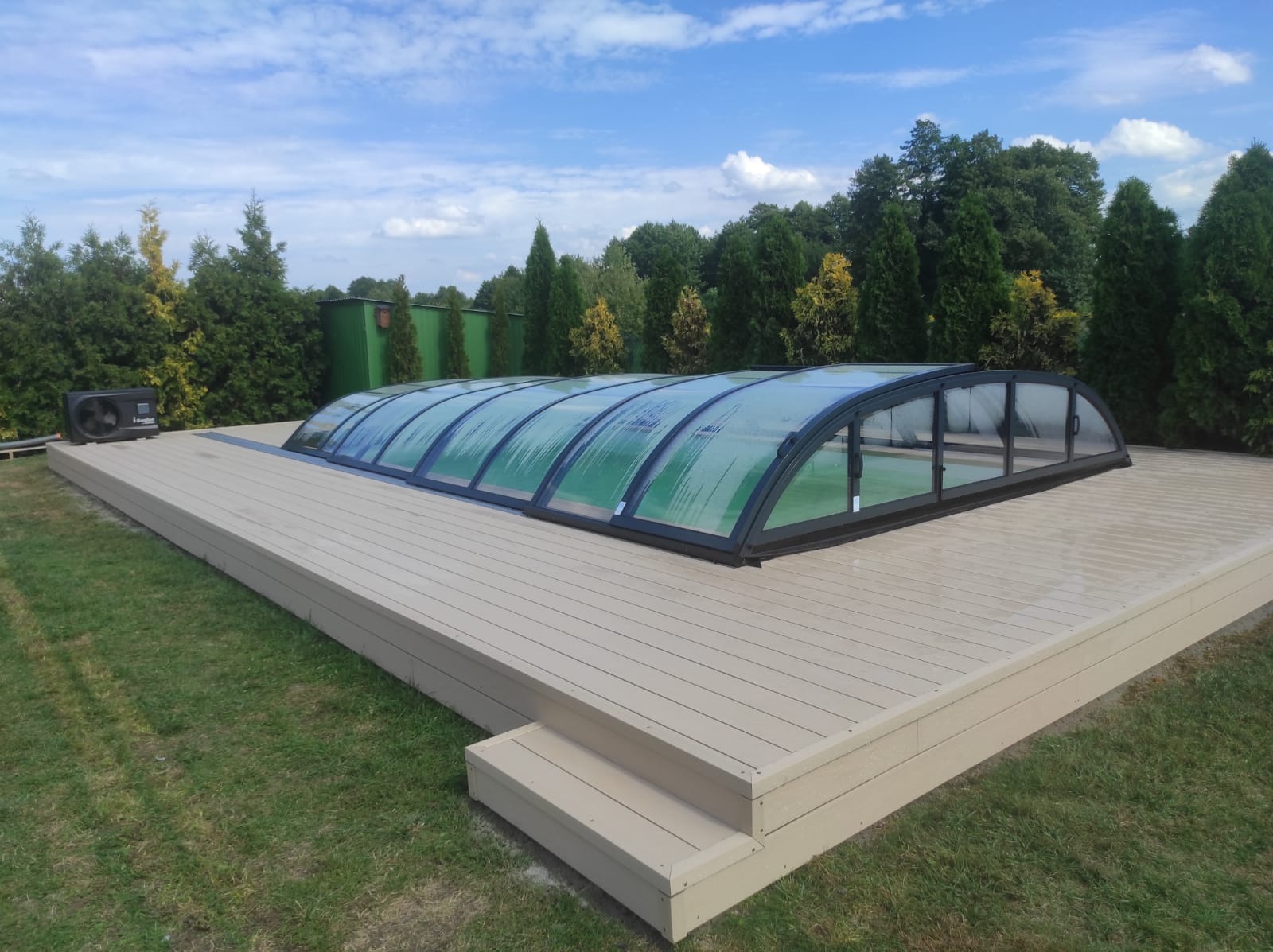 Beżowy taras z zadaszeniem basenu na tle zieleni. Widoczne schodki i pompa ciepła. Błękitne niebo z chmurami w tle.