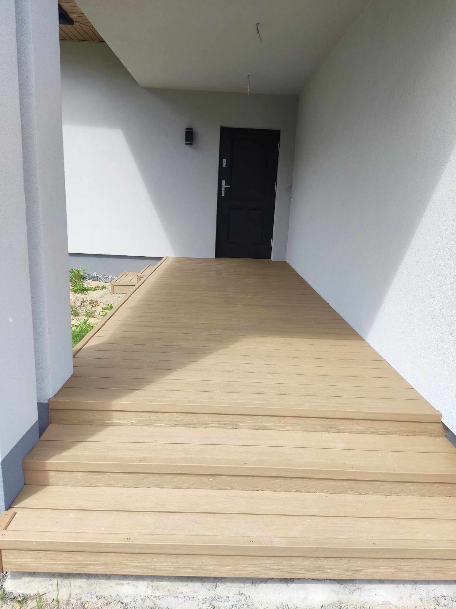 Jasny taras z desek prowadzący do drzwi wejściowych domu. Widoczne schody i fragment ogrodu z roślinnością. Ujęcie z dołu, podkreślające solidność konstrukcji.