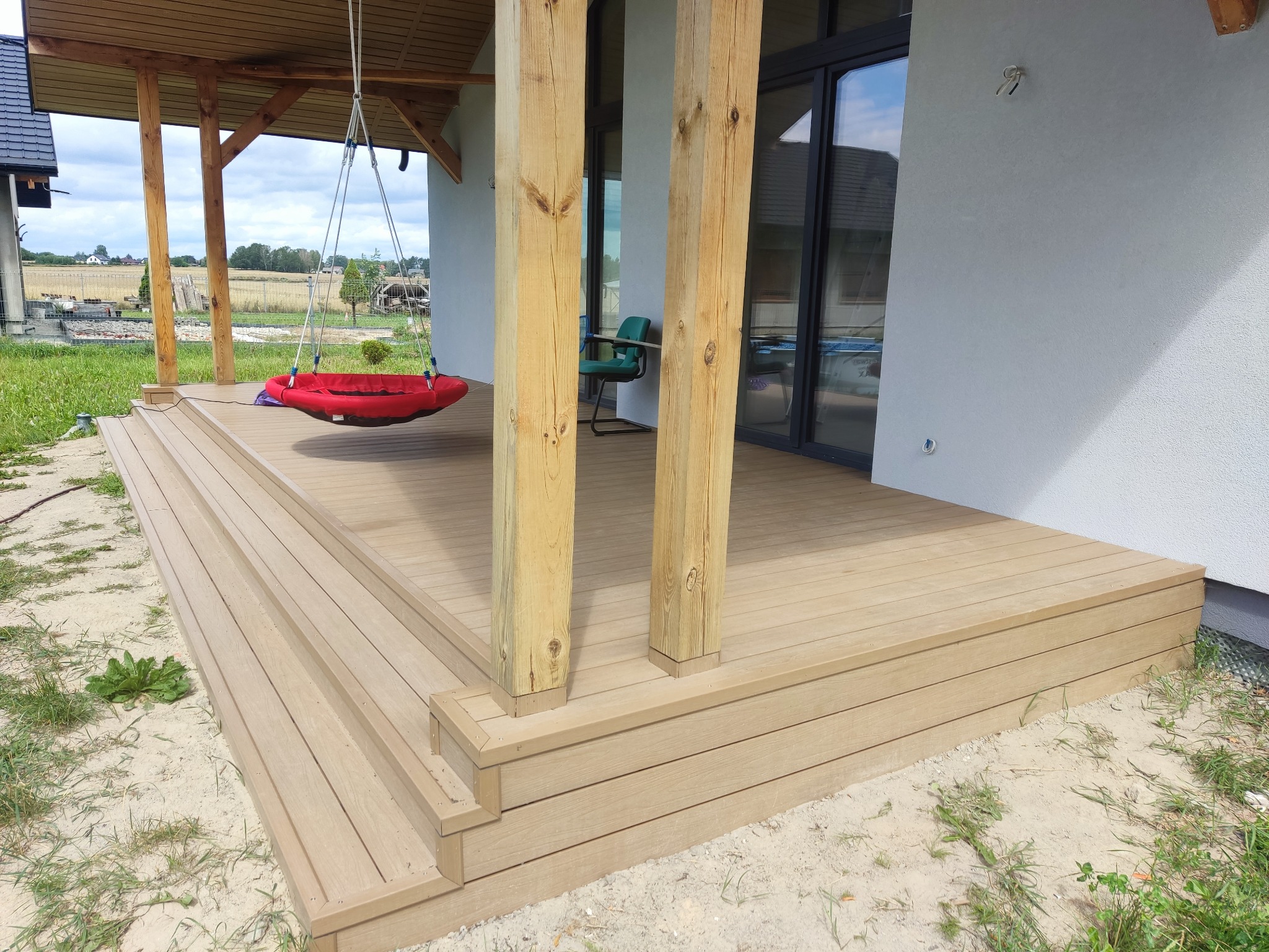 Beżowy taras z desek z huśtawką typu bocianie gniazdo. Widoczne drewniane słupy podtrzymujące zadaszenie i przeszklone drzwi przesuwne.