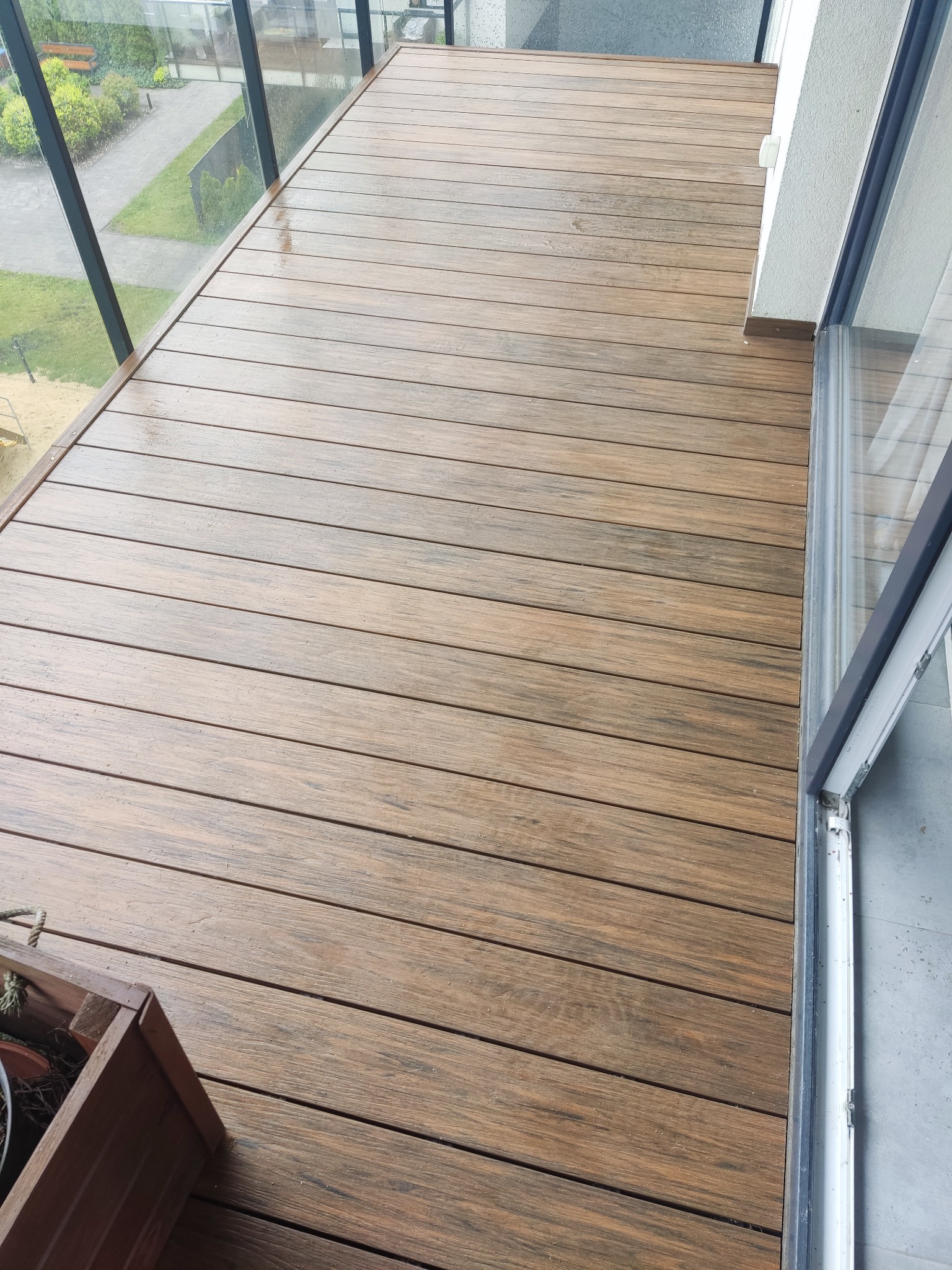 Nowoczesny balkon z desek w odcieniach brązu w Brzezinach. Szklana balustrada i drewniana donica dodają elegancji.