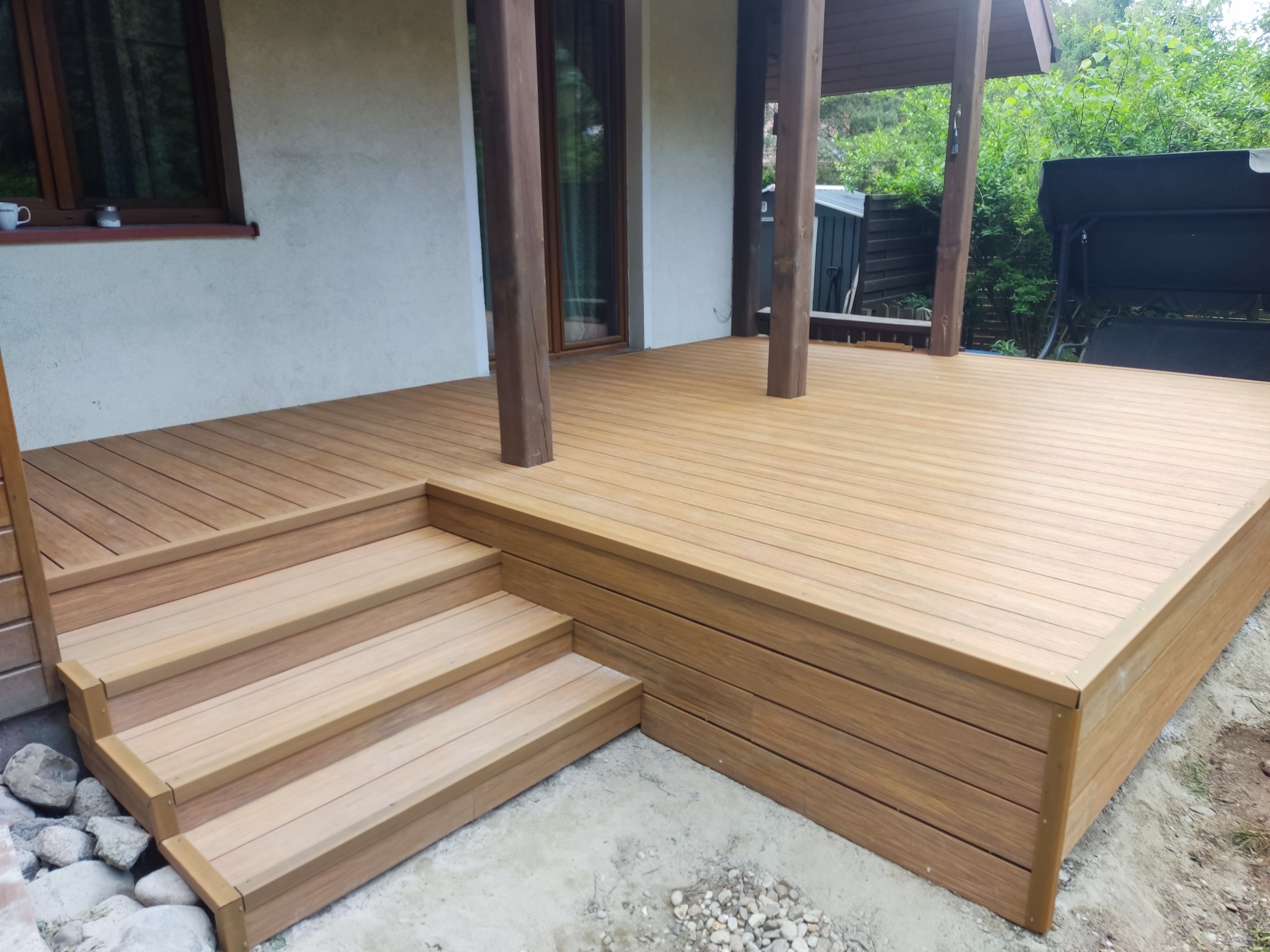 Jasny taras z brązowymi słupami wspartymi na drewnianej konstrukcji, z niskimi schodami i widocznymi kamieniami u podstawy. Widok na ogród w tle.