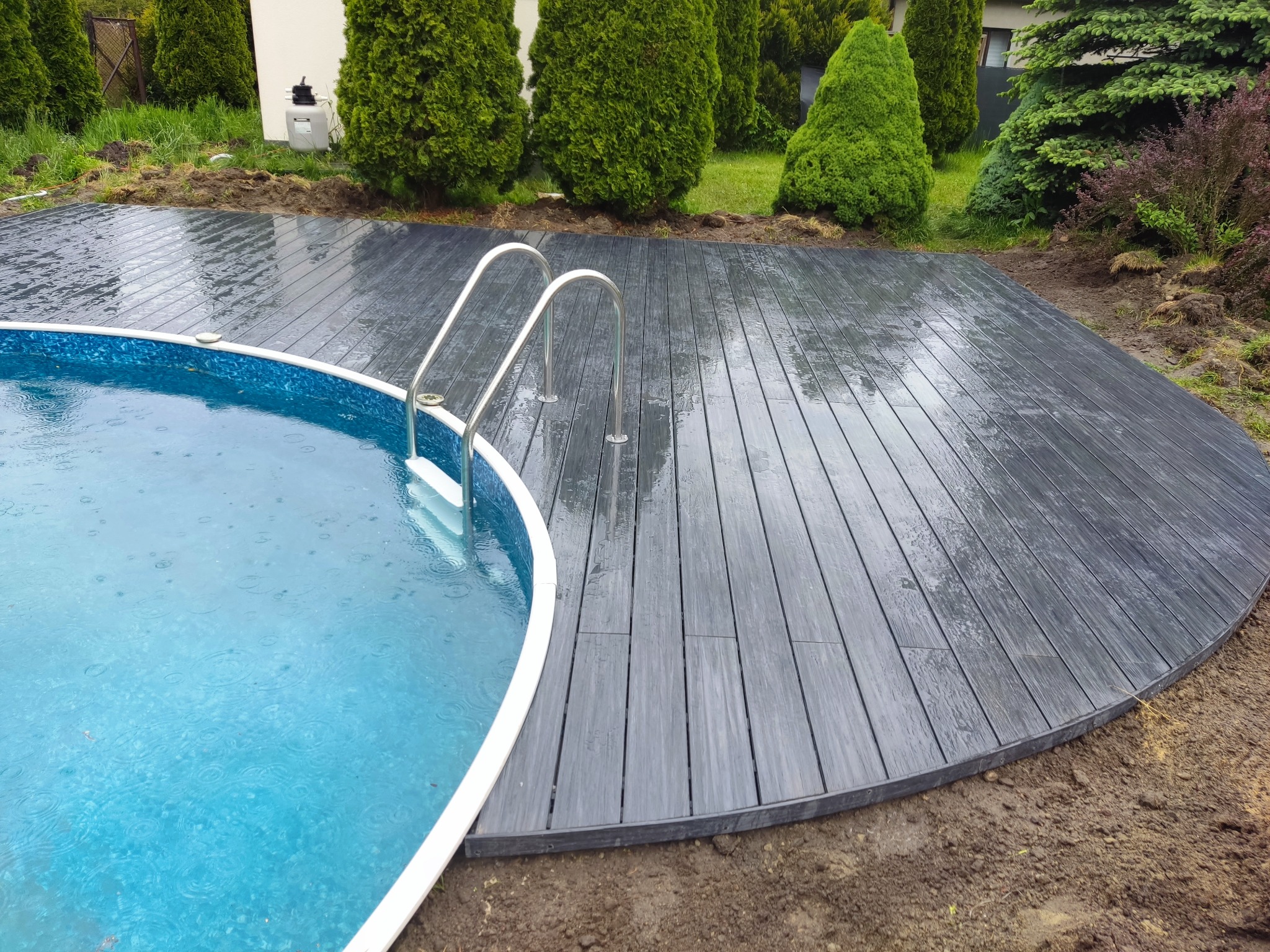 Szara, mokra nawierzchnia przy basenie z drabinką, otoczona zielenią i ziemią. Widok z góry na zaokrąglony kształt podestu.