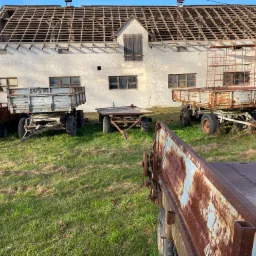 Stary budynek gospodarczy z odsłoniętą konstrukcją dachu w trakcie remontu, otoczony zardzewiałymi przyczepami rolniczymi na trawiastym terenie.