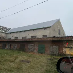 Budynek gospodarczy w Sieradzu z częściowo wymienionym poszyciem dachowym, widoczna więźba dachowa na jednej części i blaszane pokrycie na drugiej. Czerwona ceglana podmurówka i szara elewacja...