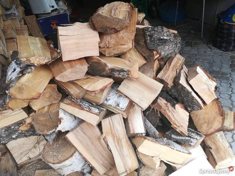 Stos pociętego drewna kominkowego z widoczną korą brzozy, ułożony na szarym bruku, w tle biały sprzęt AGD i niebieskie pudełko.