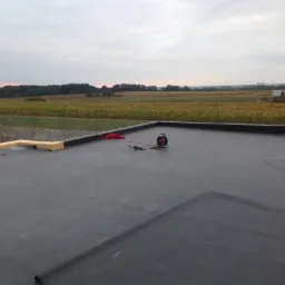 Czarny dach płaski pokryty papą termozgrzewalną w trakcie prac wykończeniowych, widoczne narzędzia dekarskie i fragment krajobrazu z polami uprawnymi w tle.