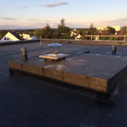 Płaski dach pokryty czarną membraną bitumiczną z widocznymi kominkami wentylacyjnymi i kominem spalinowym z metalowym daszkiem, w tle zabudowa jednorodzinna i zachodzące słońce.