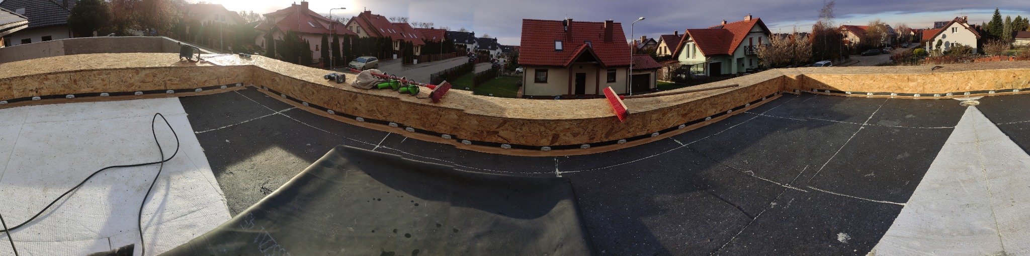 Panorama z dachu w trakcie prac: widoczna warstwa hydroizolacyjna, drewniana obudowa z płyt OSB, narzędzia dekarskie i zabudowa domów jednorodzinnych w tle.