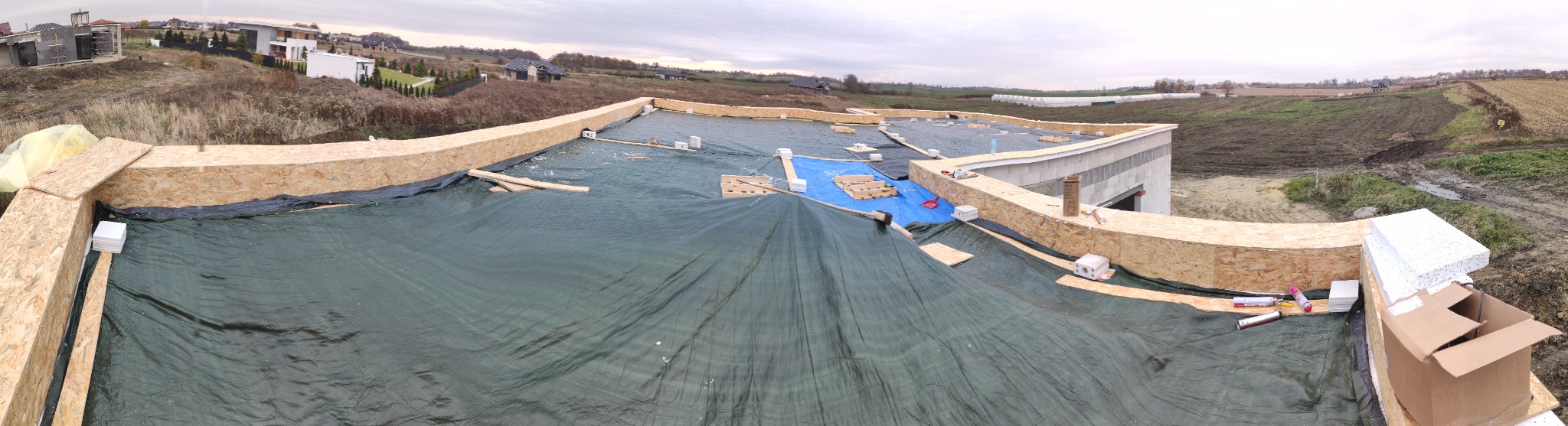 Szeroki kadr na dach płaski w trakcie budowy, zabezpieczony czarną folią i częściowo niebieską plandeką, z widocznymi elementami konstrukcyjnymi z płyt OSB i bloczków, w tle krajobraz wiejski...