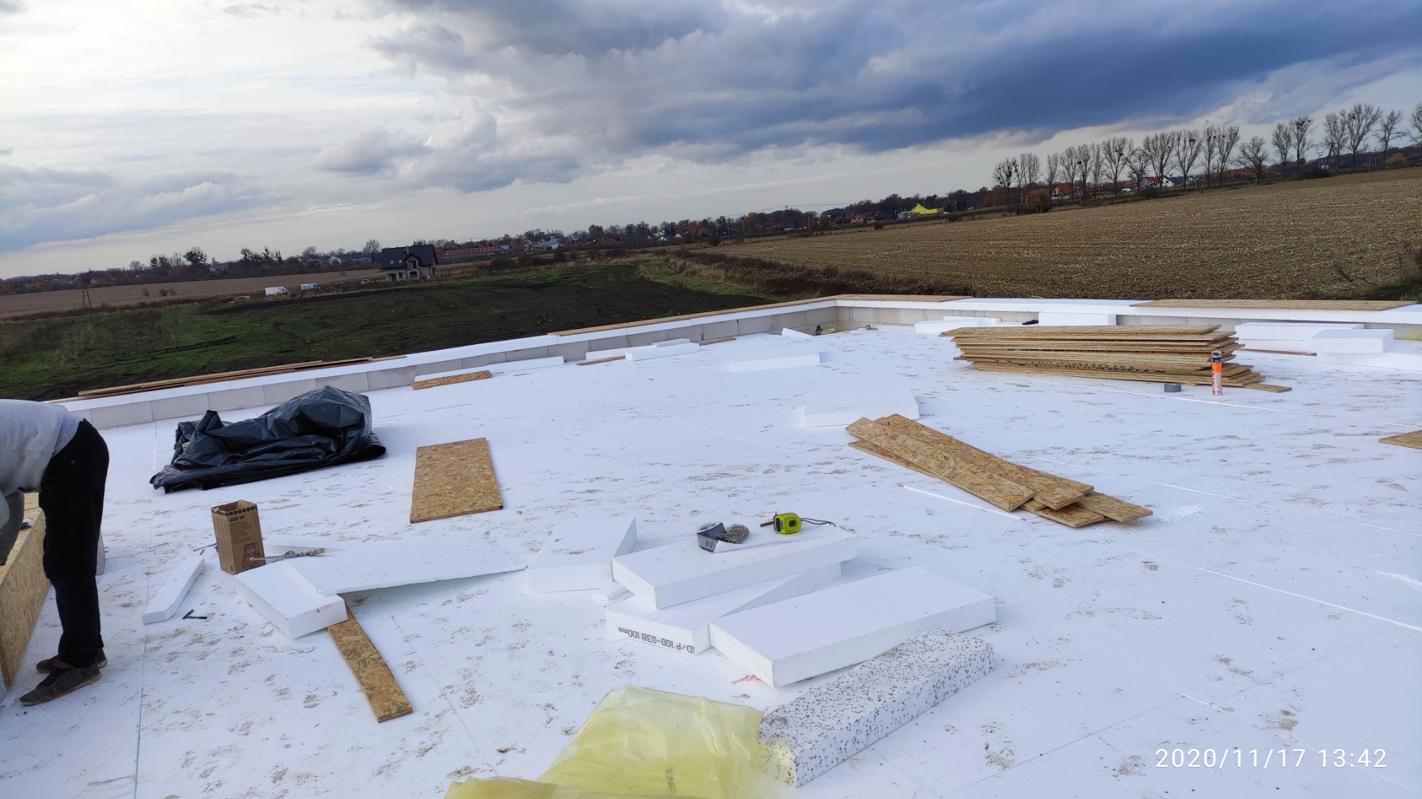 Prace na dachu: białe płyty izolacyjne, płyty OSB, narzędzia i pracownik w trakcie montażu na tle pól i pochmurnego nieba.