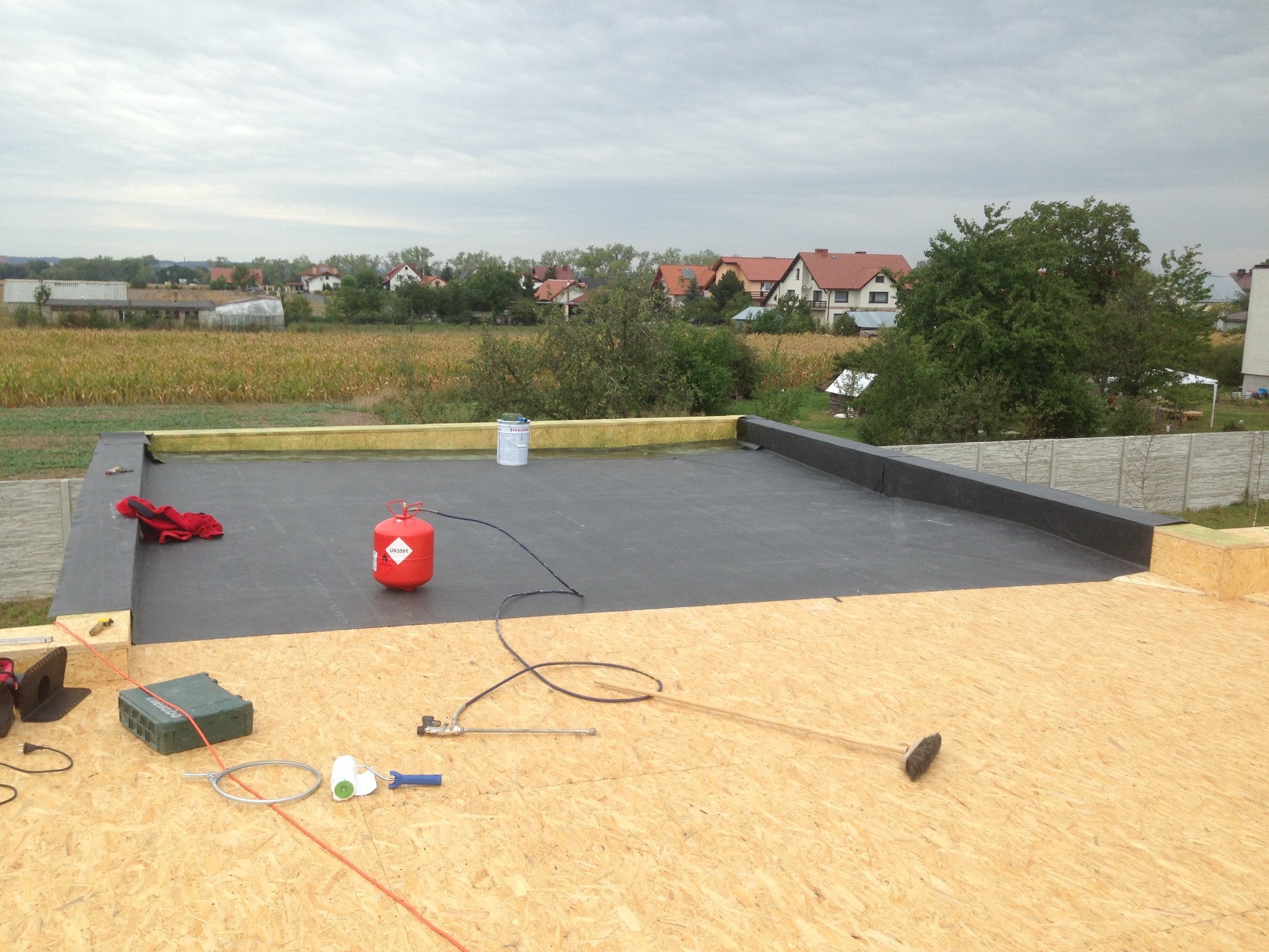 Płaski dach w trakcie uszczelniania papą termozgrzewalną, widoczna butla z gazem, narzędzia dekarskie, oraz fragment krajobrazu z domami w tle.