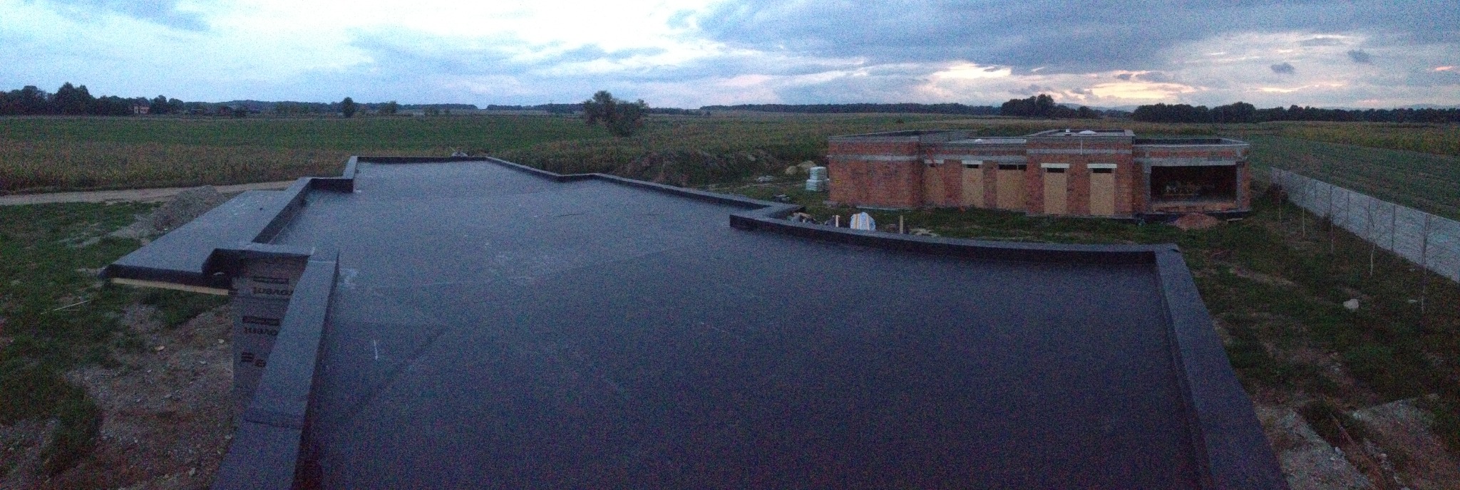 Widok z góry na czarny, płaski dach pokryty papą termozgrzewalną, z widoczną konstrukcją budynku w tle i rozległym polem w oddali pod pochmurnym niebem.