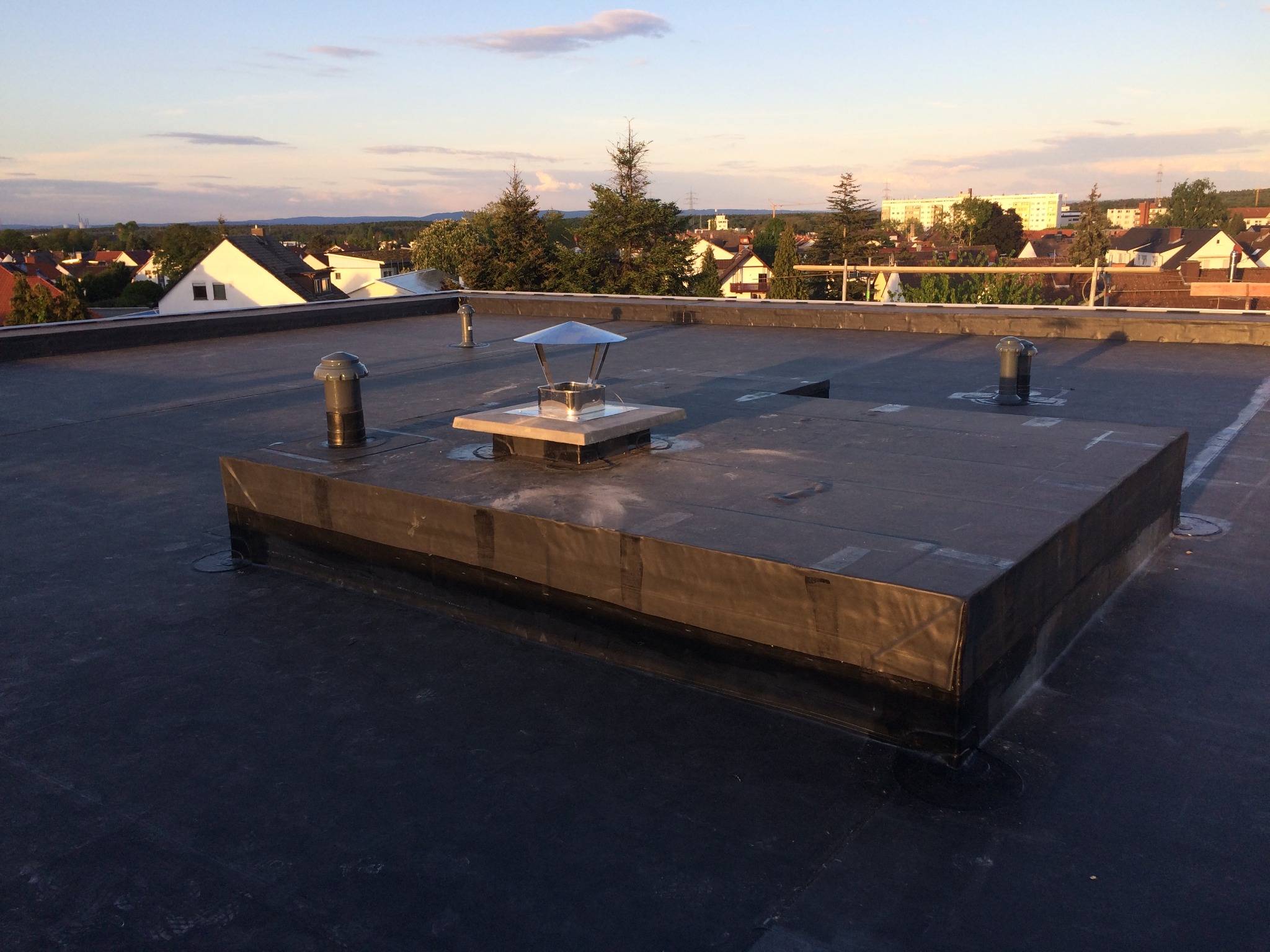 Płaski dach pokryty czarną membraną bitumiczną z widocznymi kominkami wentylacyjnymi i kominem spalinowym z metalowym daszkiem, w tle zabudowa jednorodzinna i zachodzące słońce.