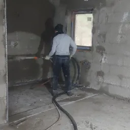 Wnętrze w trakcie prac wykończeniowych: pracownik aplikuje tynk na ścianę przy pomocy węża, widoczne przewody elektryczne na suficie pokrytym pianą izolacyjną oraz okno z pomarańczową ramą.