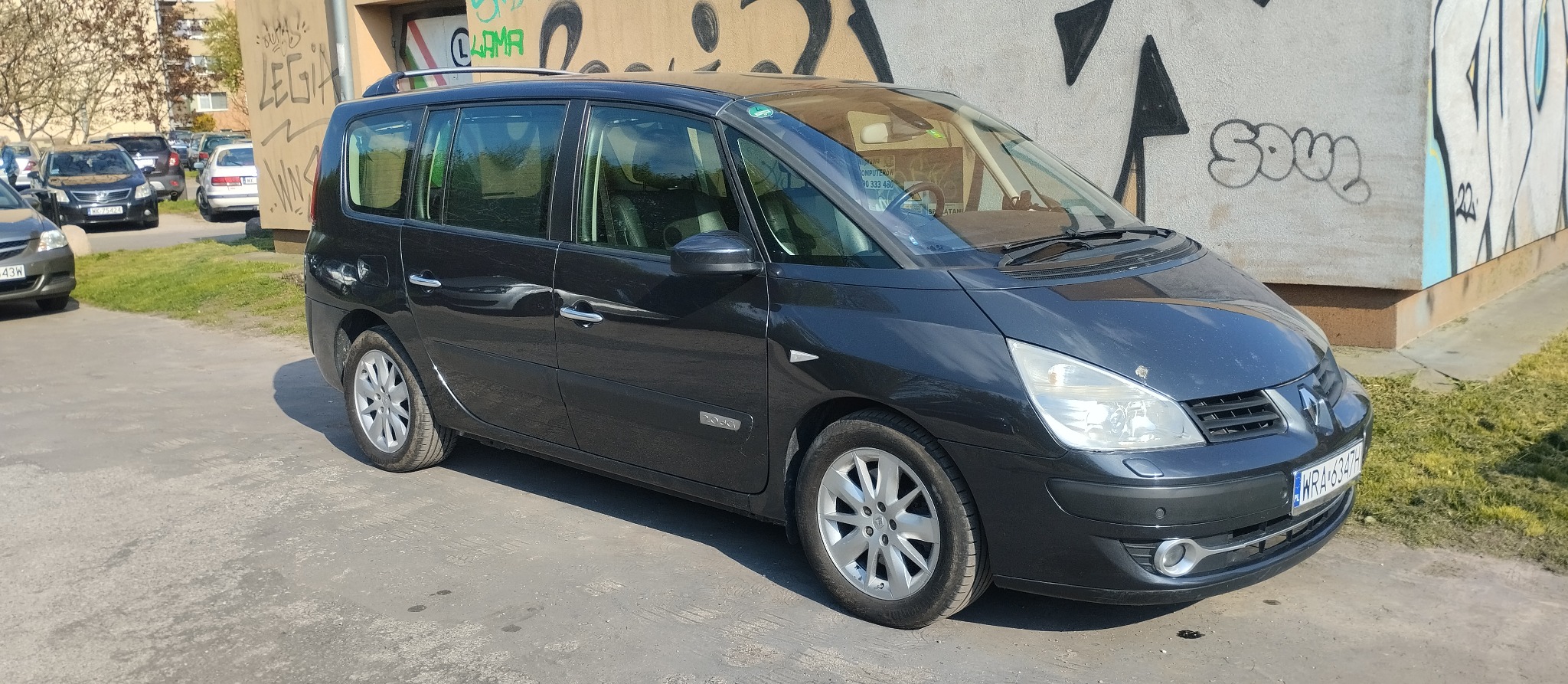 Czarny minivan Renault Espace zaparkowany na betonowym placu, z graffiti na ścianie w tle. Widoczny numer rejestracyjny WRA 6347H.
