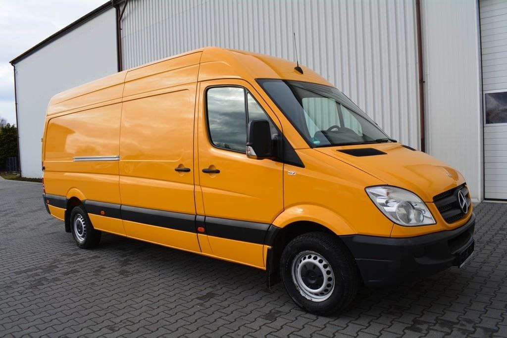 Żółty bus marki Mercedes-Benz Sprinter, zaparkowany na brukowanej powierzchni przed magazynem z blachy falistej.