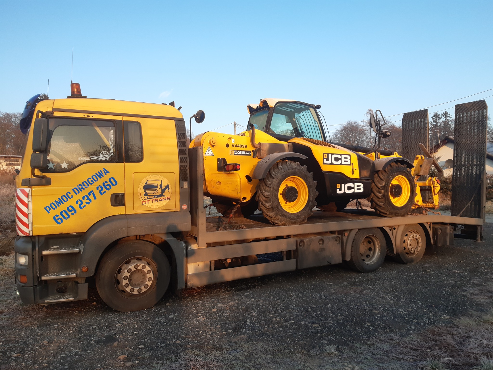 Żółta laweta z numerem telefonu pomocy drogowej przewozi żółtą ładowarkę teleskopową JCB, zabezpieczoną łańcuchami, na tle zimowego krajobrazu.