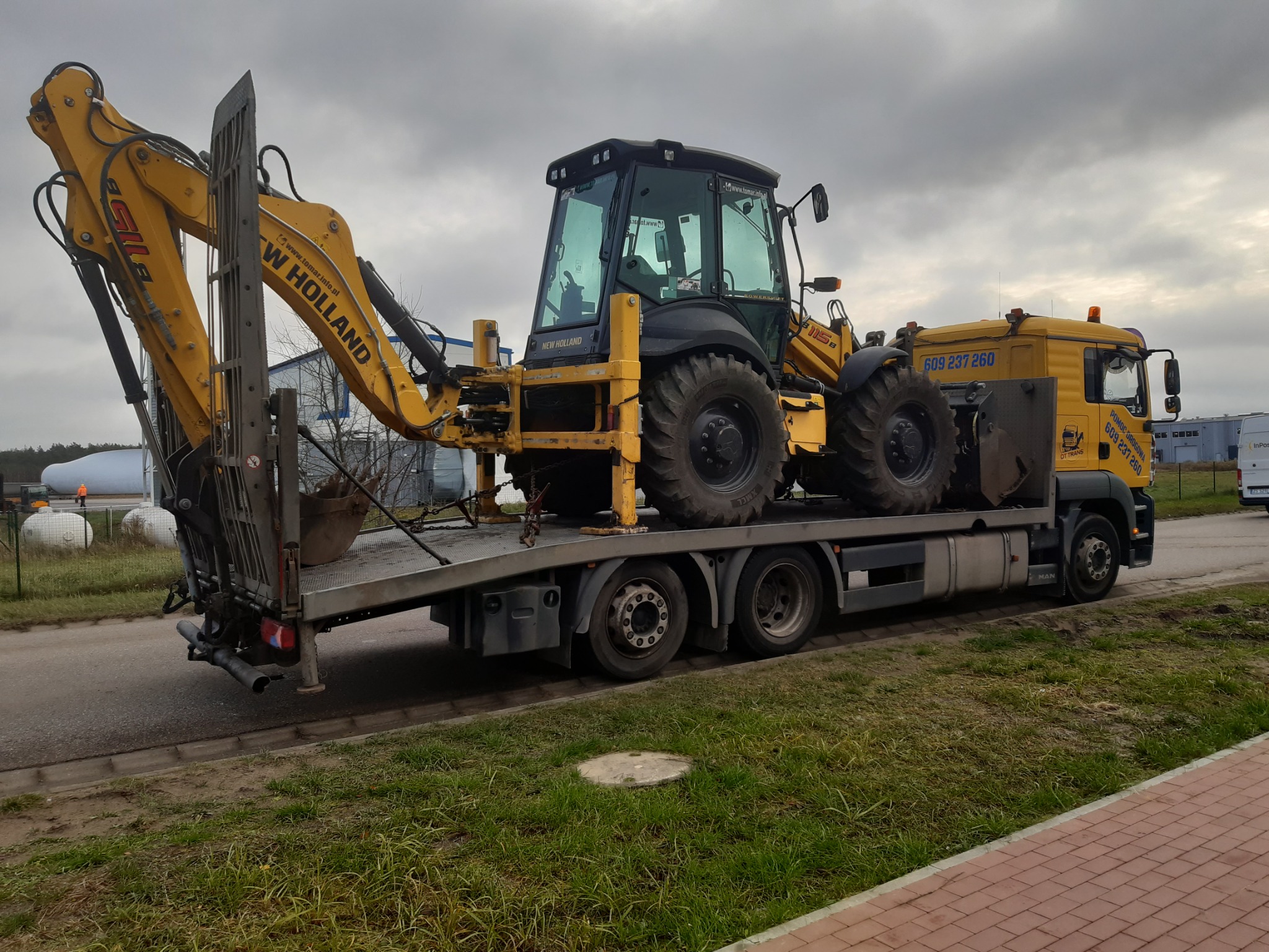 Żółta koparko-ładowarka New Holland 115B przewożona na platformie lawety MAN z widocznym numerem telefonu alarmowego i logo firmy OTT TRANS.