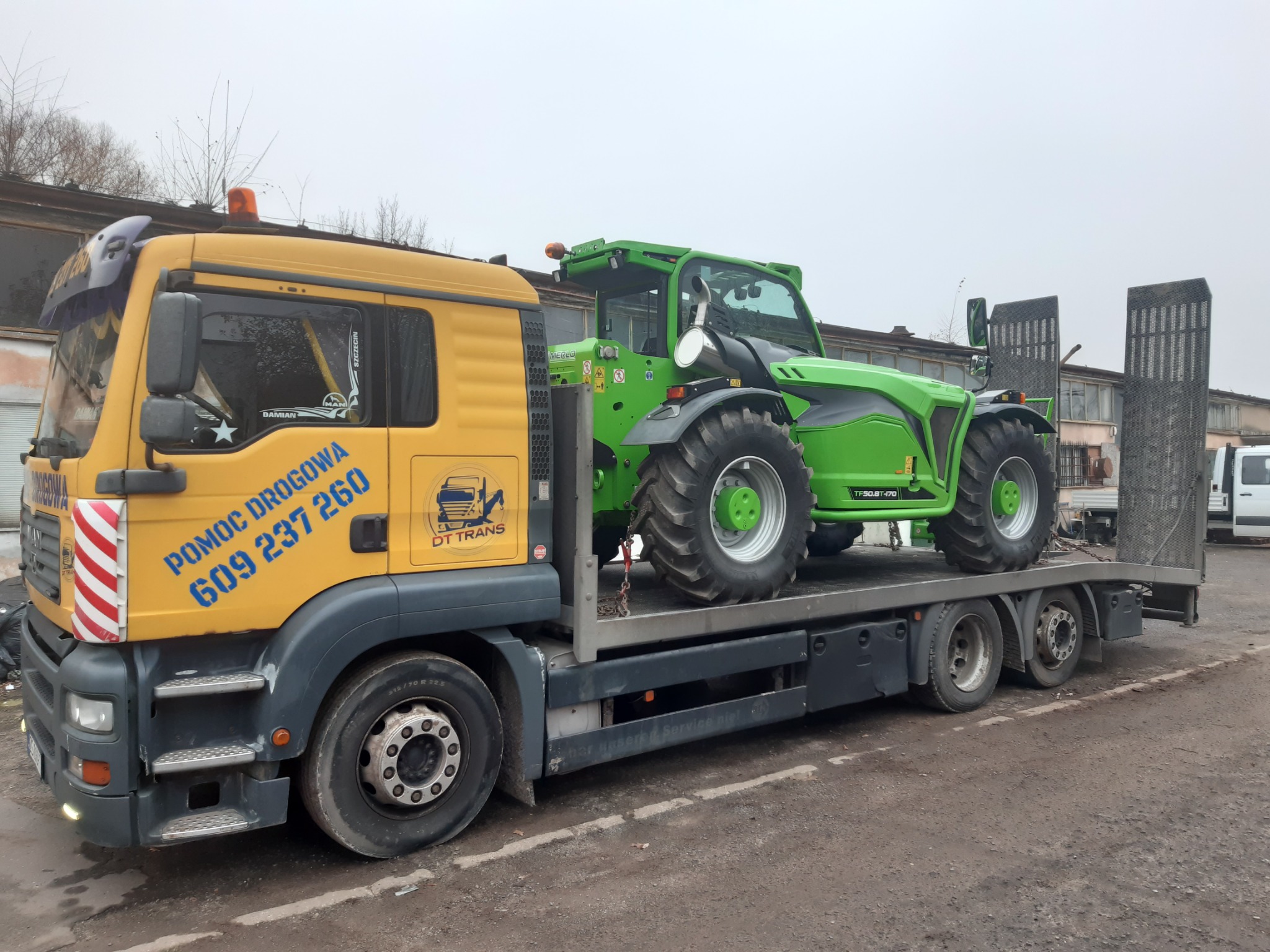 Żółta ciężarówka z napisem 'Pomoc Drogowa' i numerem telefonu przewozi na platformie zieloną ładowarkę teleskopową Merlo TF50.8T-170 na tle budynku przemysłowego.