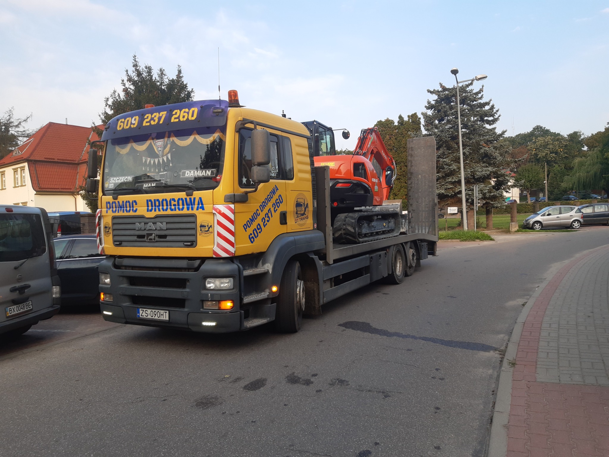 Żółta ciężarówka z napisem 'Pomoc Drogowa' i numerem telefonu, przewożąca pomarańczową mini-koparkę na platformie, zaparkowana na skrzyżowaniu w mieście.