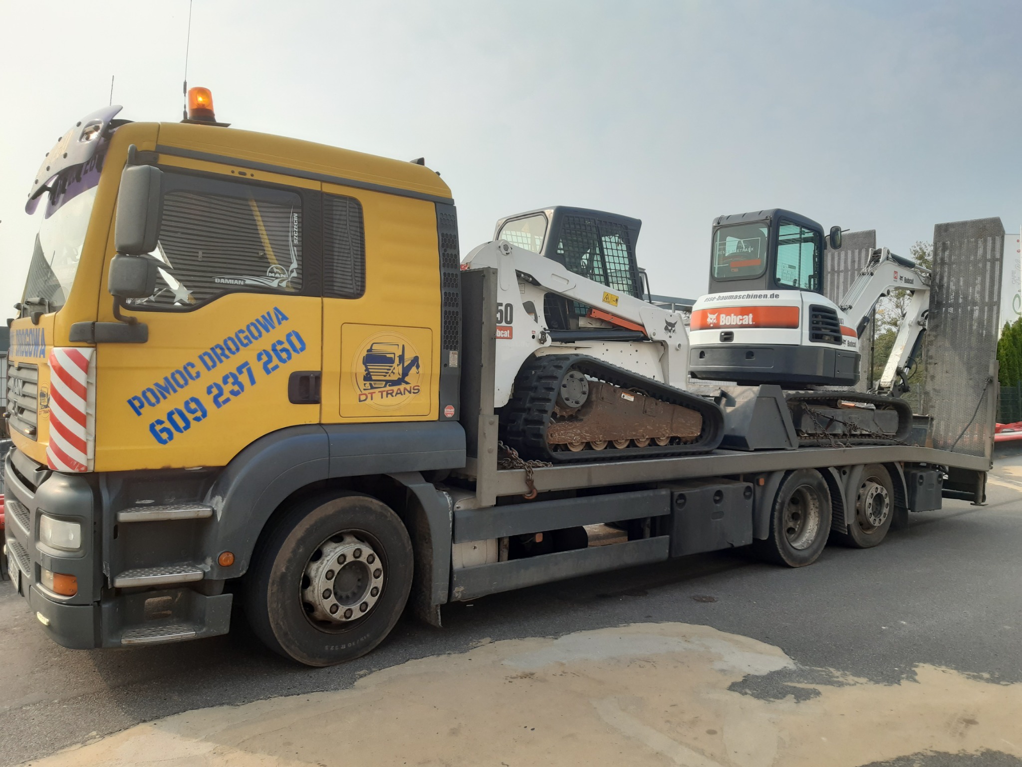 Żółta laweta z napisem 'Pomoc Drogowa 609 237 260 DT Trans' przewożąca dwa białe minikoparki Bobcat na platformie, widok z boku na tle szarego nieba.