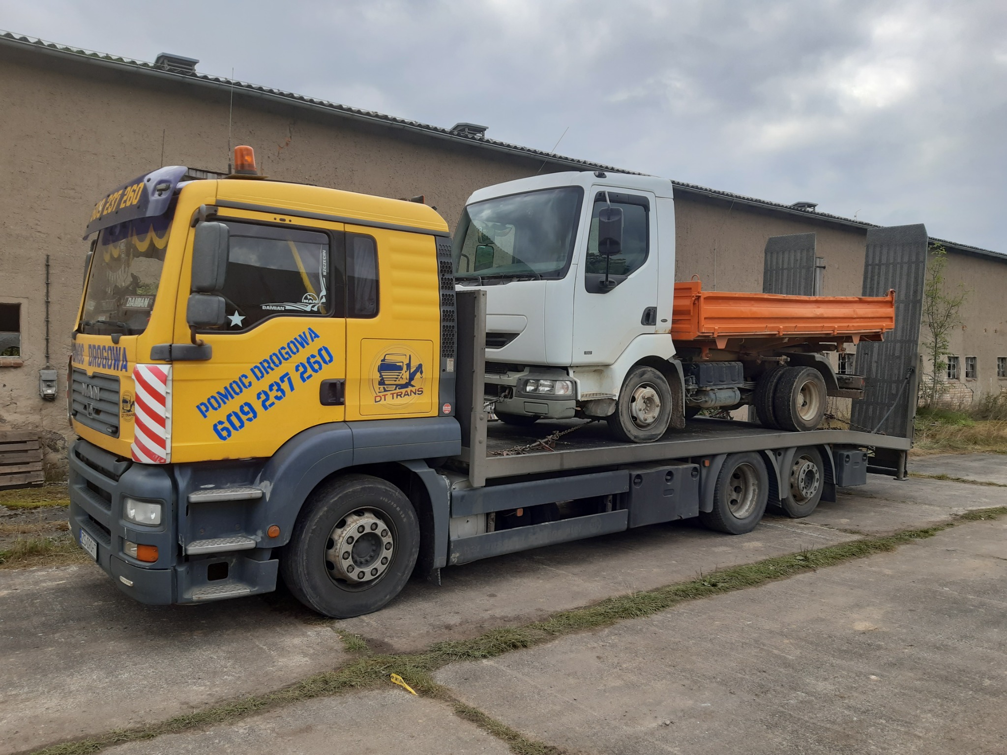 Żółta ciężarówka pomocy drogowej MAN z biało-czerwoną szrafurą na kabinie przewozi na platformie białą wywrotkę Renault z pomarańczową skrzynią ładunkową na betonowym placu przed budynkiem.