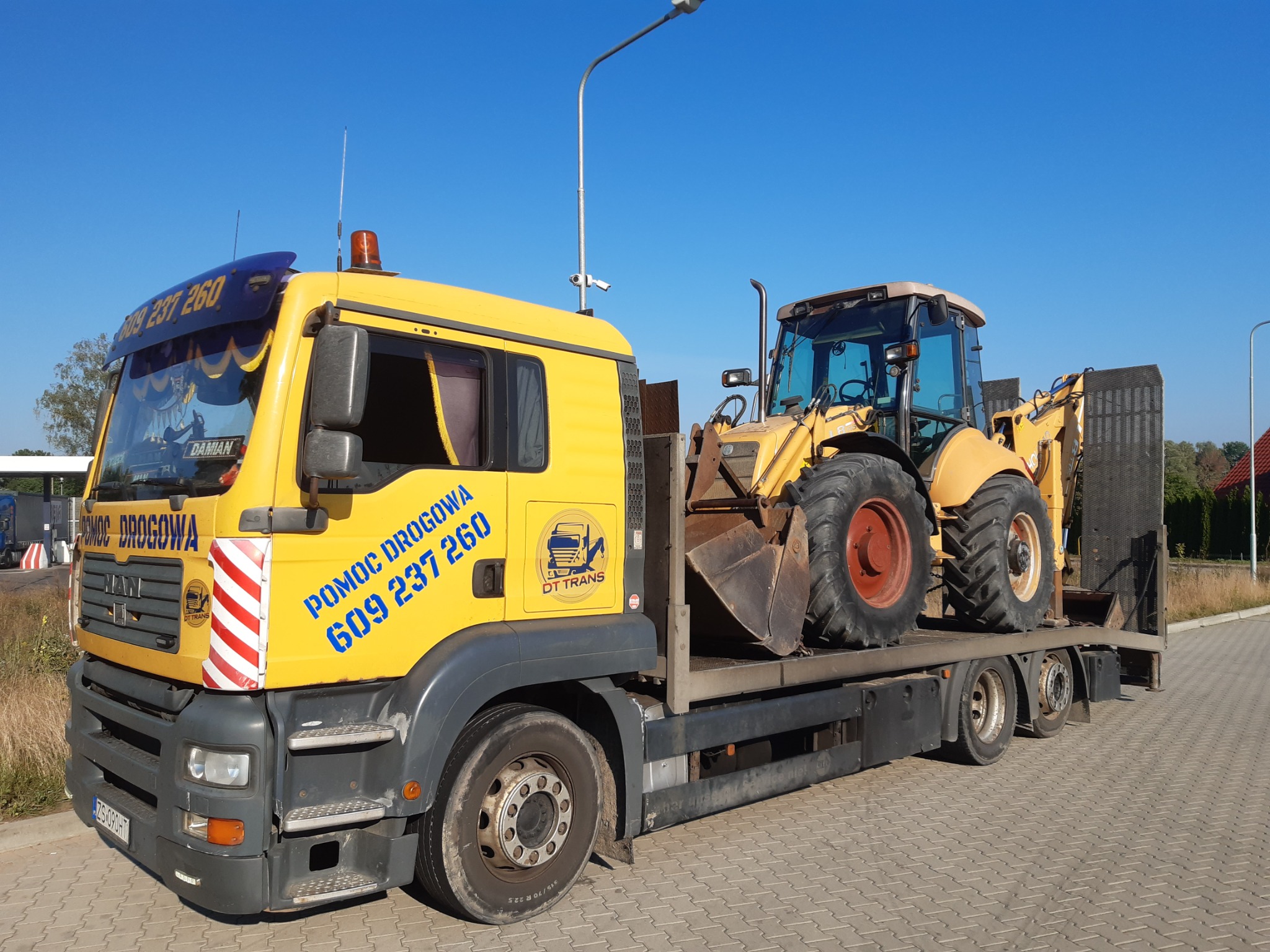 Żółta ciężarówka pomocy drogowej MAN z naklejkami i numerem telefonu, przewożąca na platformie żółtą koparko-ładowarkę na tle błękitnego nieba.