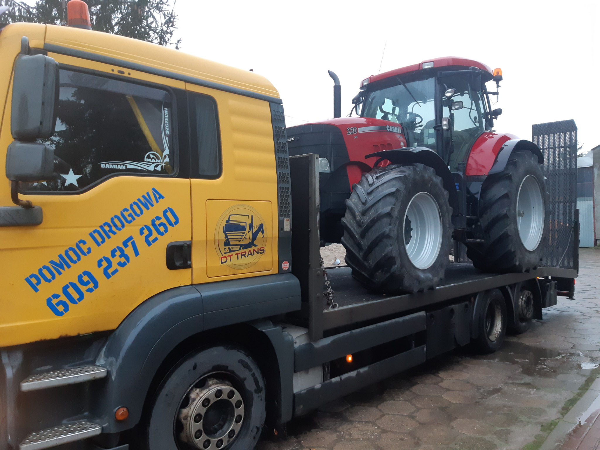 Żółta ciężarówka z niebieskim napisem 'Pomoc Drogowa' przewozi czerwony traktor marki Case na platformie.