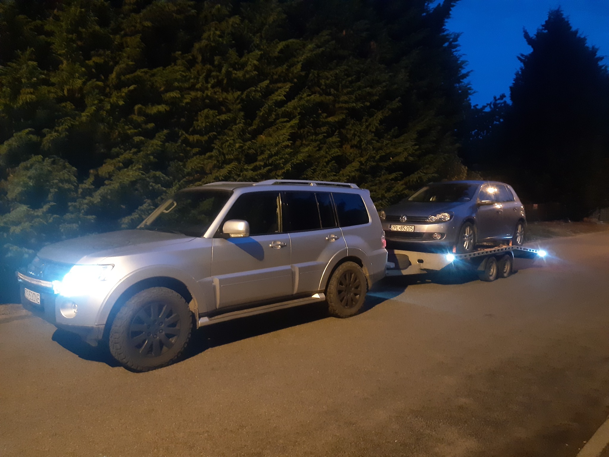 Srebrny SUV holuje na przyczepie autolawety srebrnego Volkswagena Golfa, nocne ujęcie na tle drzew.