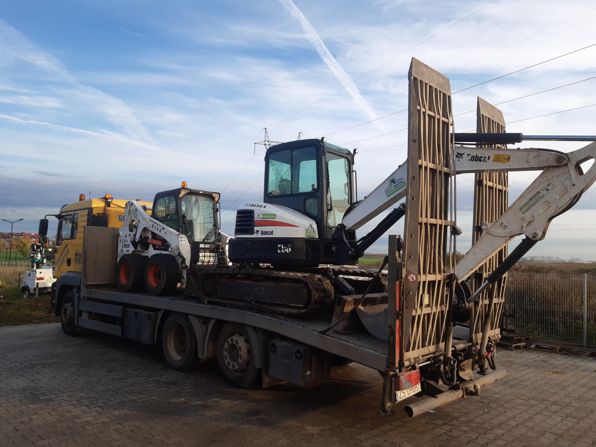 Żółta ciężarówka z lawetą przewożąca dwie maszyny budowlane Bobcat na tle błękitnego nieba z smugami kondensacyjnymi