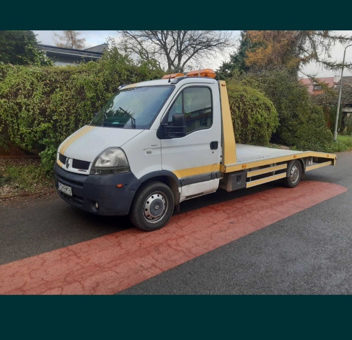 Biała laweta Renault Master z żółtymi pasami na karoserii, zaparkowana na mokrej, asfaltowej drodze z czerwonym pasem dla pieszych.