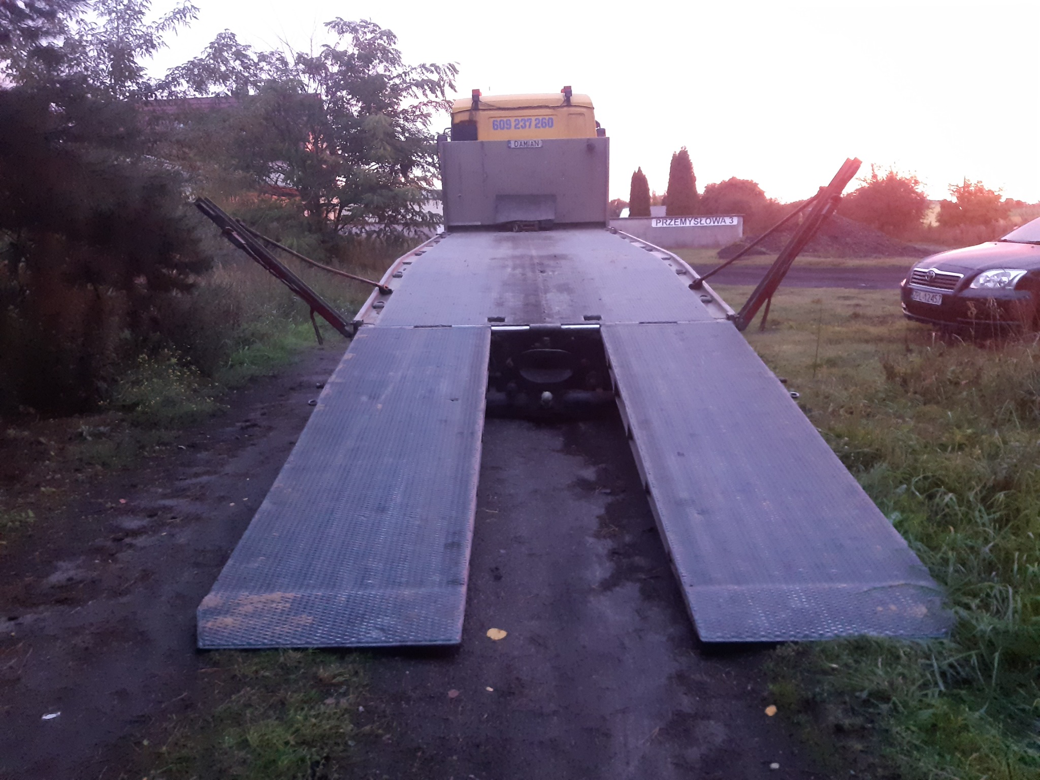 Pusta platforma lawety z rozłożonymi najazdami, widok z tyłu, na tle zieleni i zaparkowanego samochodu osobowego.