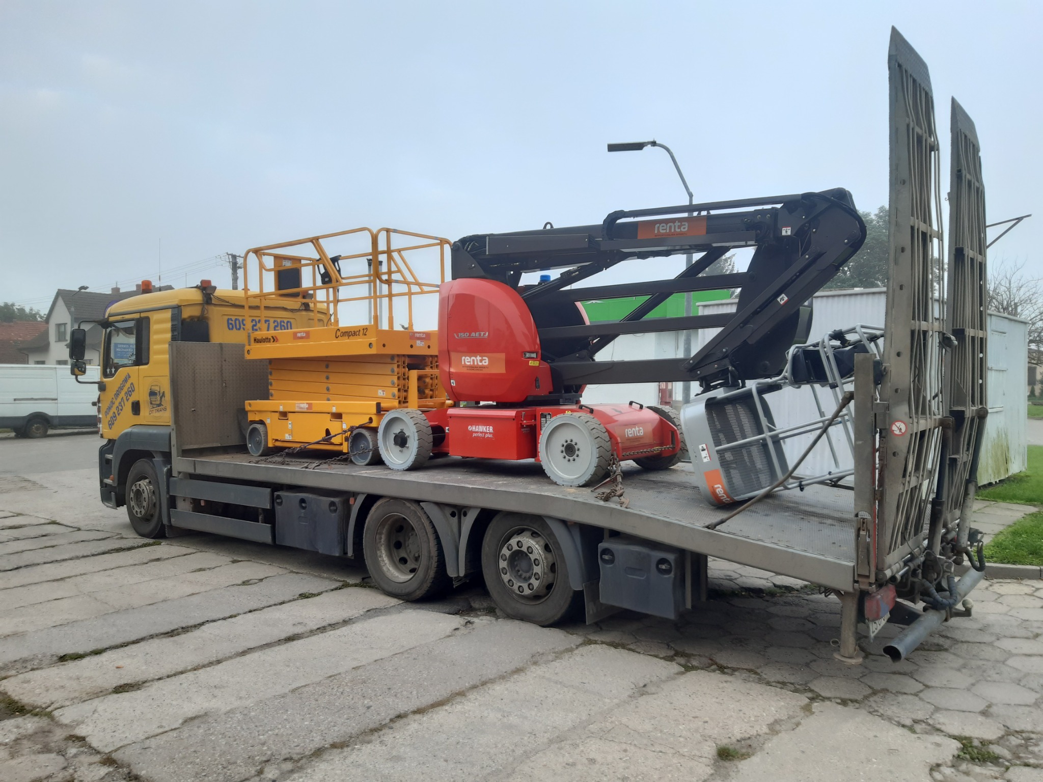 Żółta ciężarówka z platformą załadunkową przewożąca dwie podnośniki koszowe: żółty nożycowy i czerwony teleskopowy z logo 'renta', zabezpieczone łańcuchami.