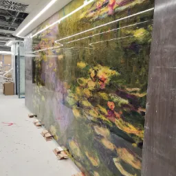 Montaż szklanej ściany z nadrukiem inspirowanym obrazem Moneta, zabezpieczonej kartonowymi podkładkami, w surowym wnętrzu z widoczną instalacją sufitową.