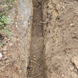 Wolf-Bud usługi minikoparki Kordian Karkocha - Wąski wykop w ziemi, prawdopodobnie pod instalacje, z widocznymi śladami pracy i nierównymi ścianami. Ziemia w kolorze brązowym, otoczenie z roślinnością i elementami betonowymi.