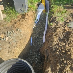 Wolf-Bud usługi minikoparki Kordian Karkocha - Wykopany rów z żółtą rurą drenażową, obsypany kamieniami i zabezpieczony białą geowłókniną. W rogu widoczny czarny zbiornik. Prace ziemne w ogrodzie.