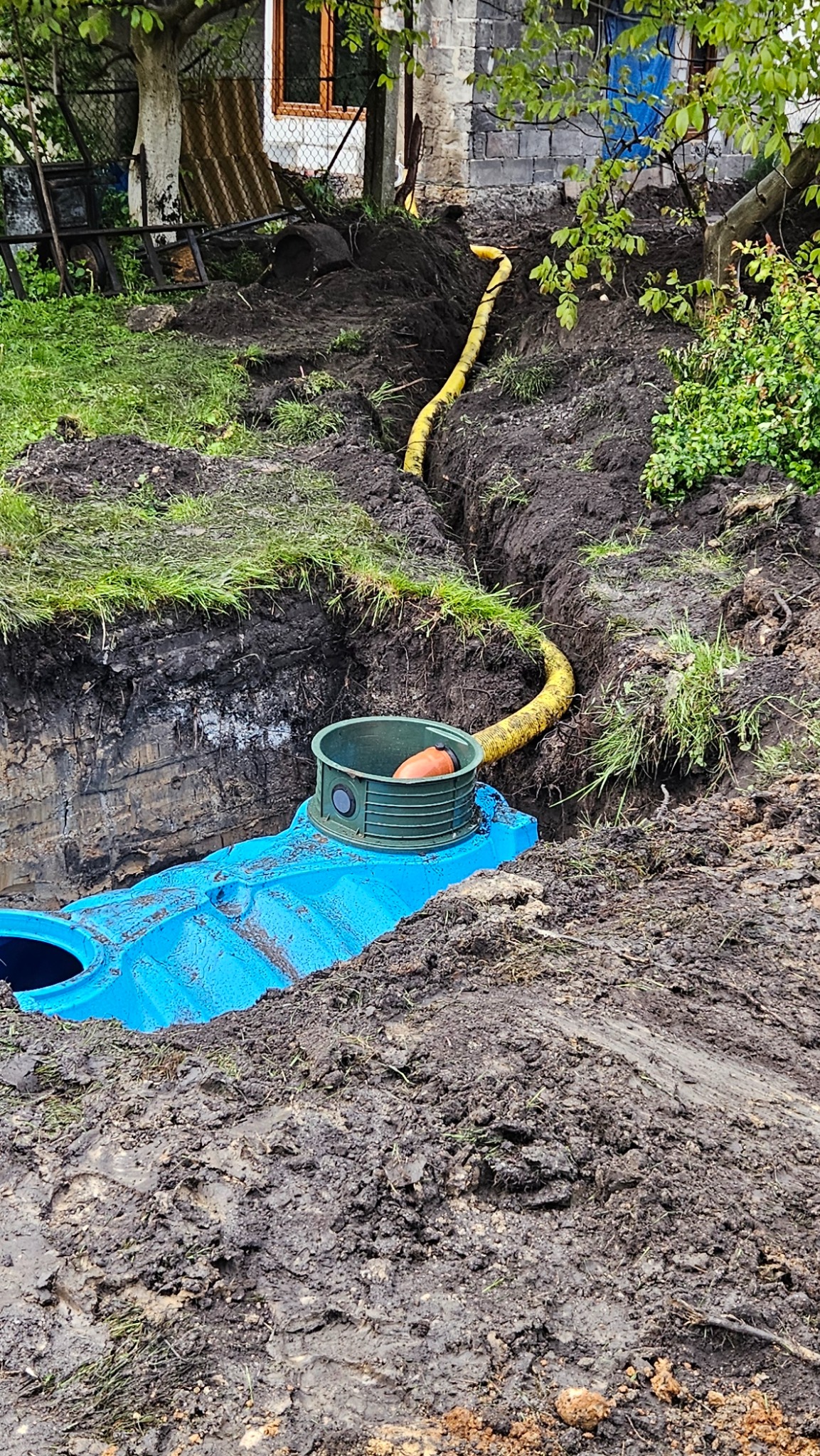 Błękitny zbiornik retencyjny w wykopie, z żółtą rurą doprowadzającą wodę. Widoczne ślady prac ziemnych i zielona trawa na skraju wykopu. Prace ziemne w toku.