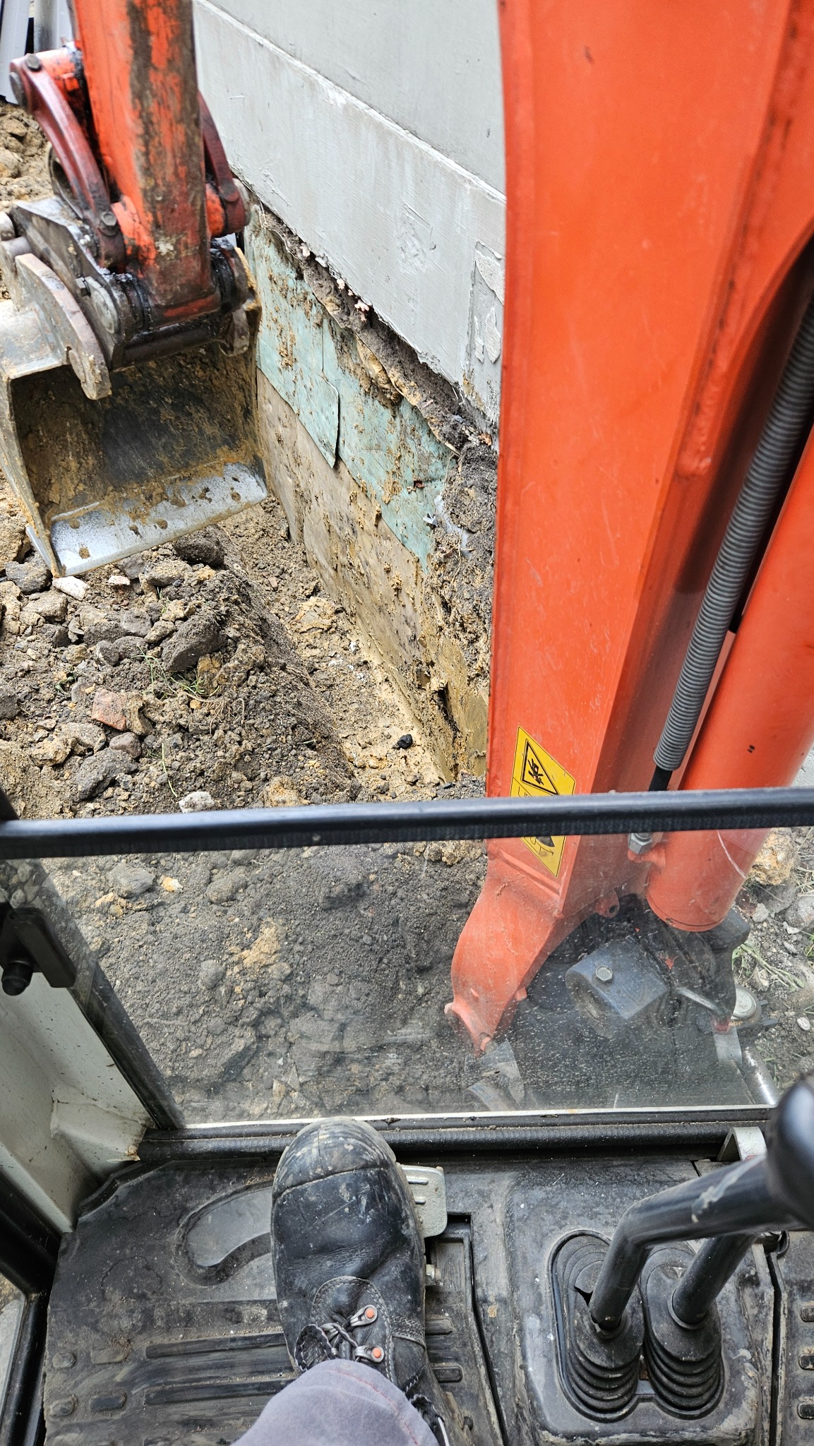 Widok z kabiny pomarańczowej koparki podczas wykopu wąskiego rowu przy fundamencie budynku. Widoczny fragment buta operatora i dźwignie sterujące.
